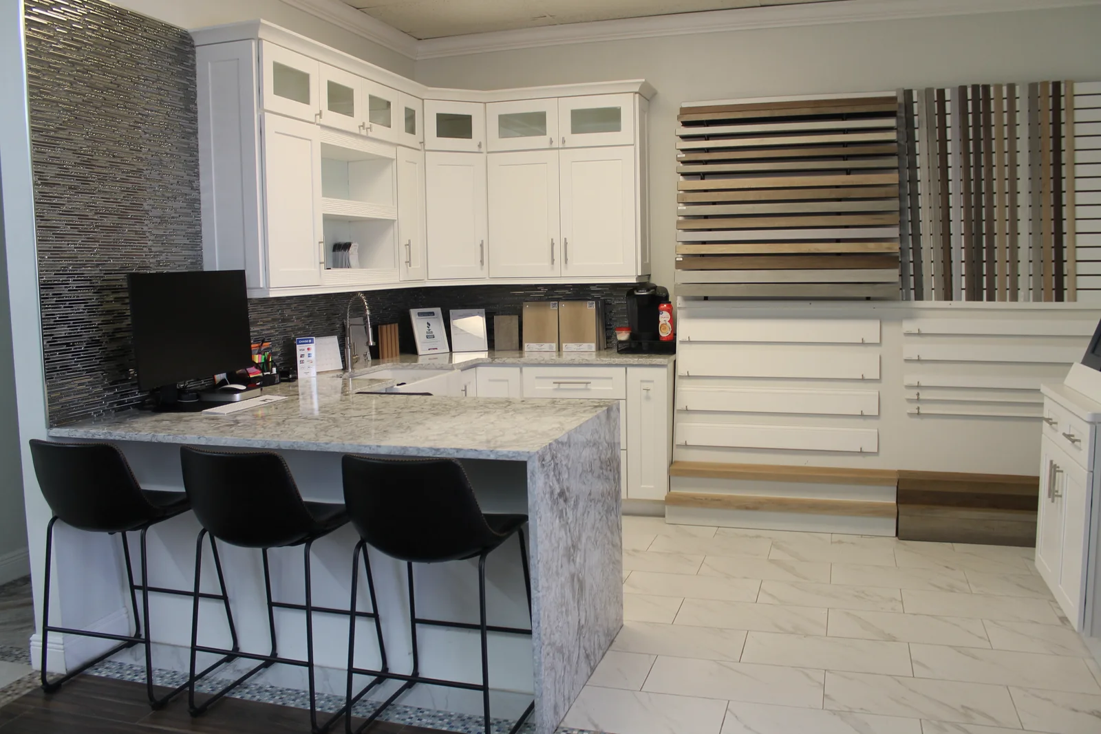 Inside the Style Home Remodeling showroom in Fort Myers — a display kitchen with a marble waterfall island beside wall racks of full-length Style SPC plank samples